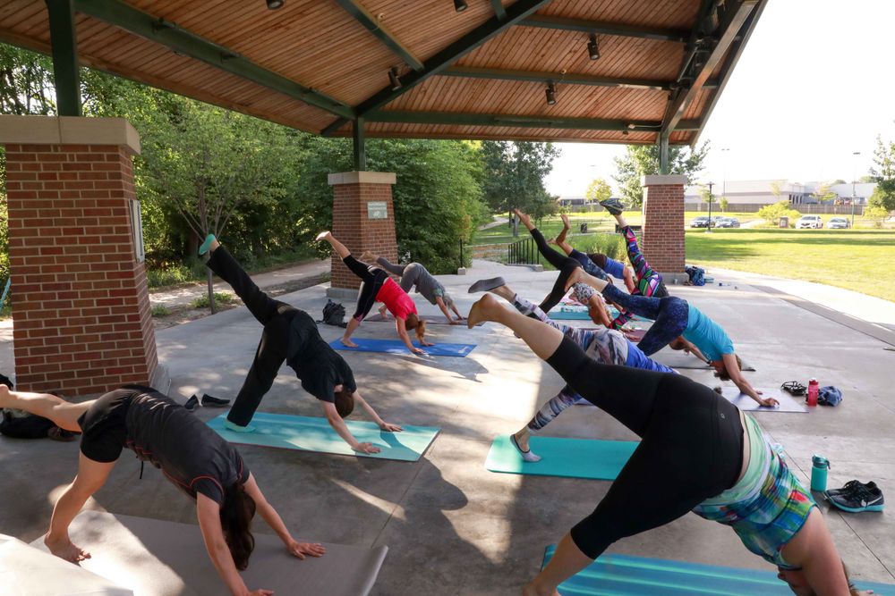 Group of people performing yoga exercises under an outdoor pavilion with brick columns and tiled roof.
