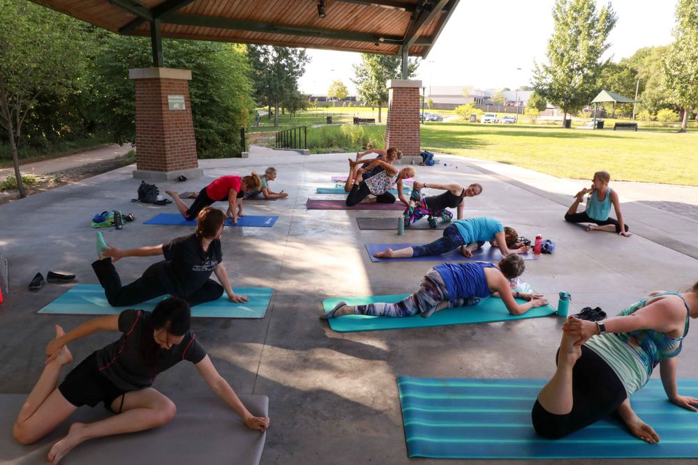 A group of people practicing yoga poses on blue mats under an outdoor pavilion during a fitness class.