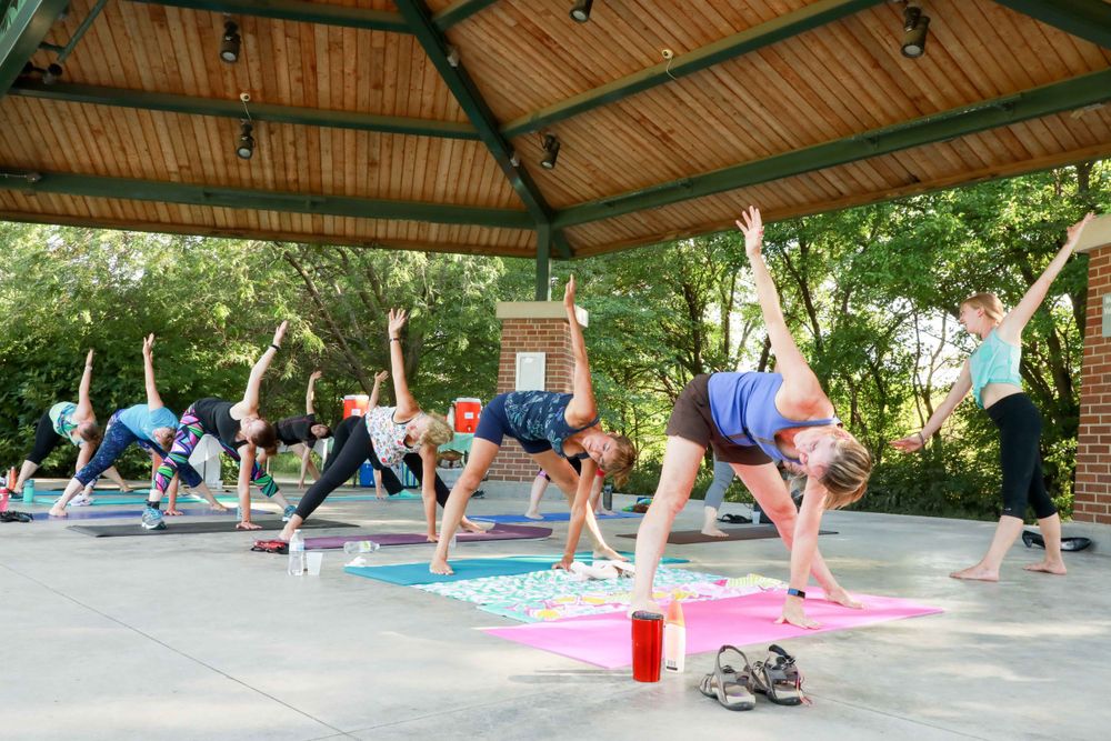 Outdoor yoga class stretches in side plank pose under a wooden pavilion with colorful yoga mats on concrete floor.