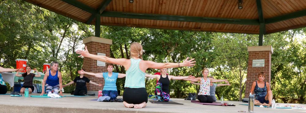 A fitness instructor leads a group exercise class under a wooden gazebo in a park setting.