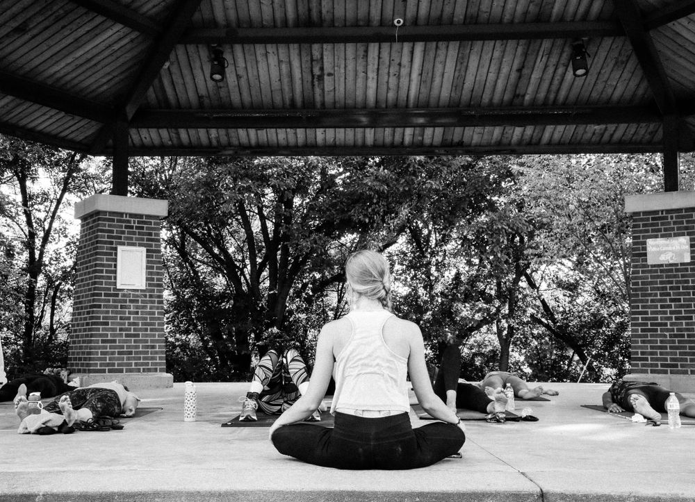 A person in white sits in lotus position leading a meditation class under an outdoor pavilion in black and white photography.