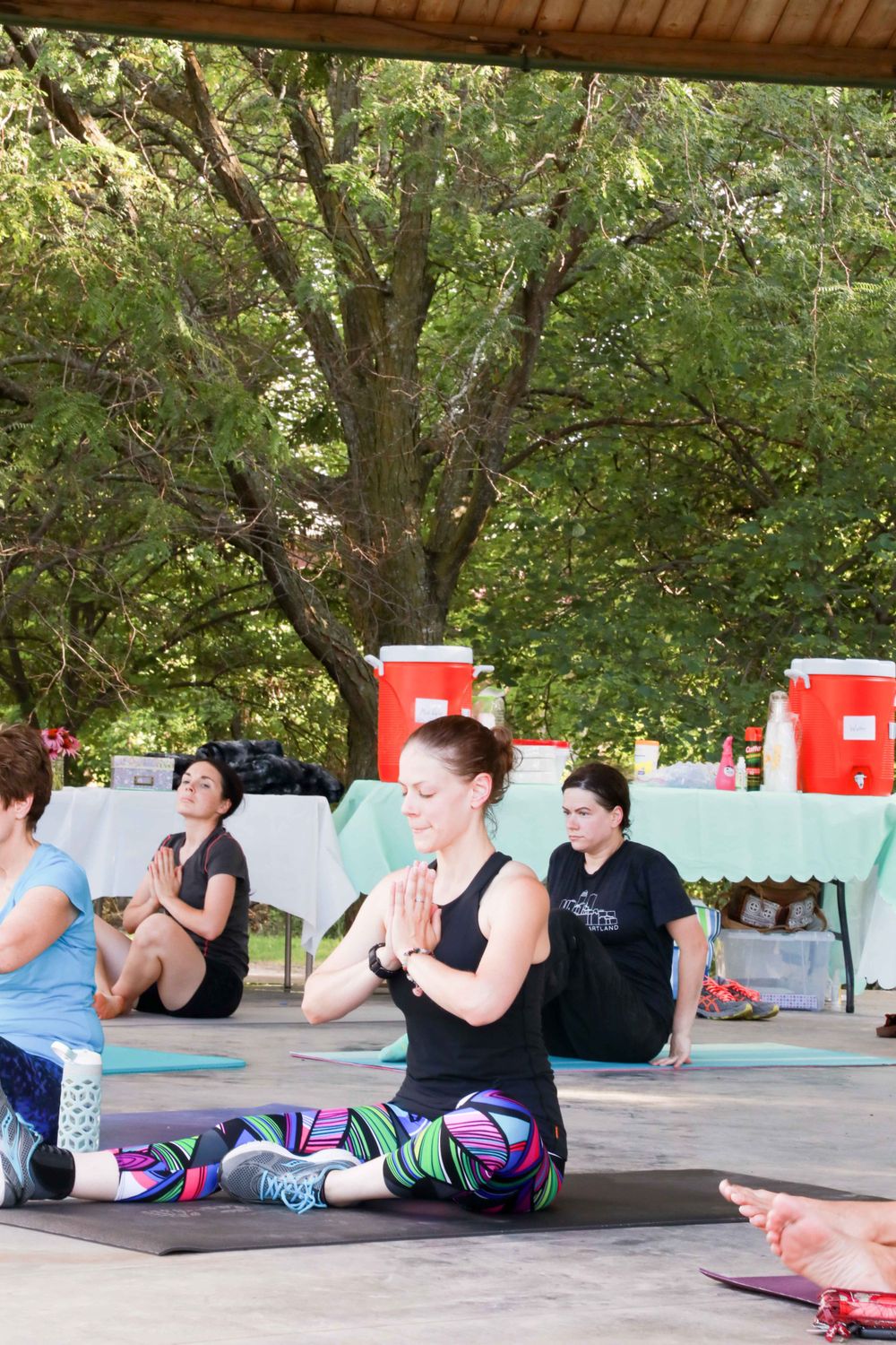 A group of people practicing yoga outdoors on mats under a covered pavilion with trees in the background.