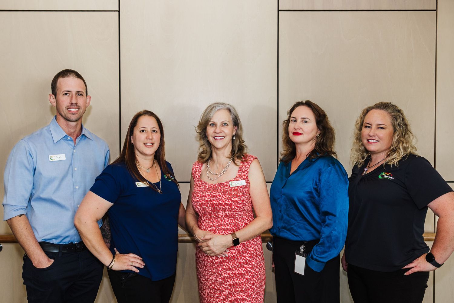 Five professionals stand together smiling in front of a beige wall wearing business attire in an office setting.