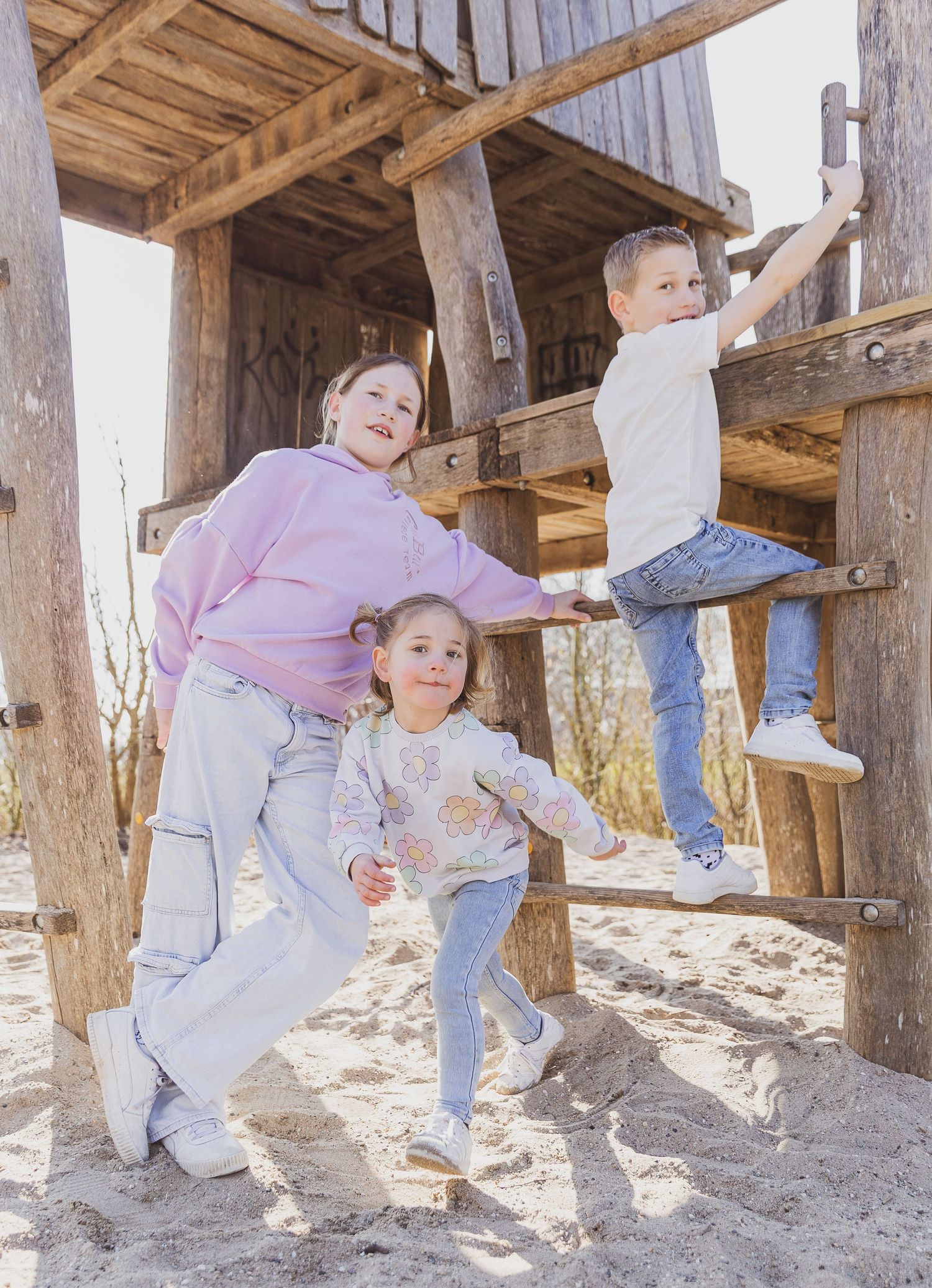 Three people playing and posing on a wooden climbing structure at a playground. Gezin op de foto fotograaf.