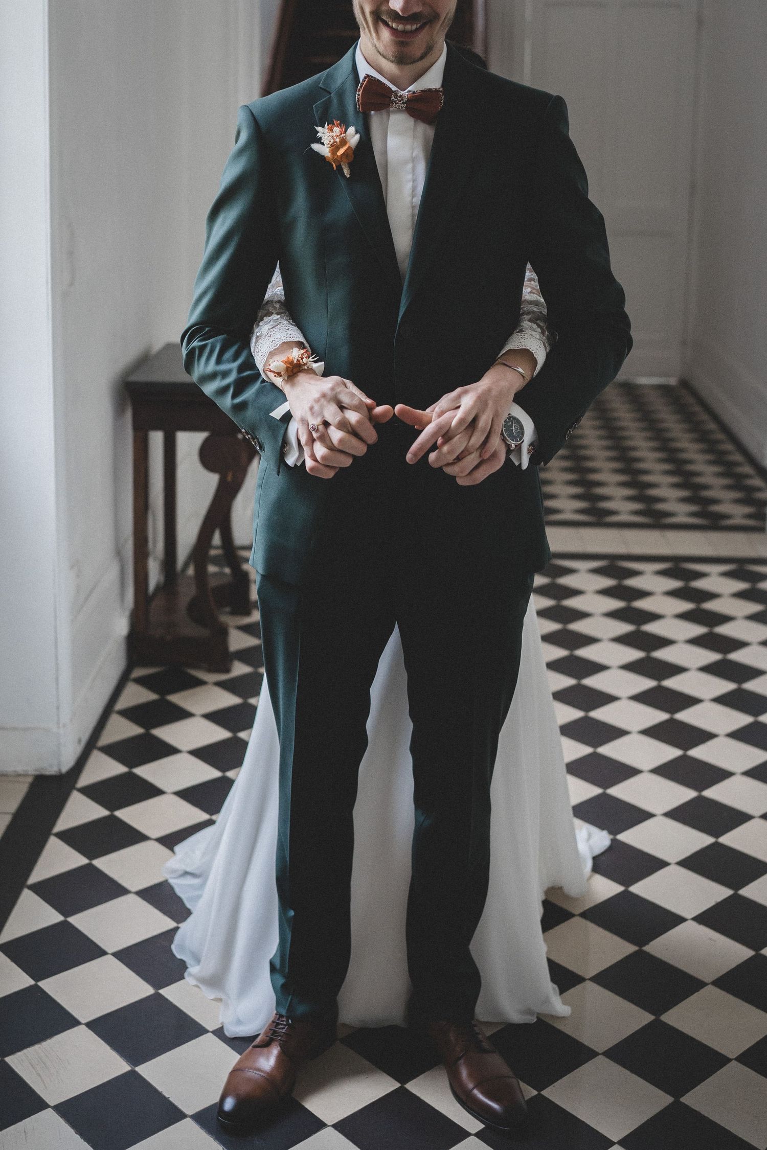 Un marié en costume vert foncé et nœud papillon se tient debout sur un sol en damier, les mains jointes pour le moment du first look.