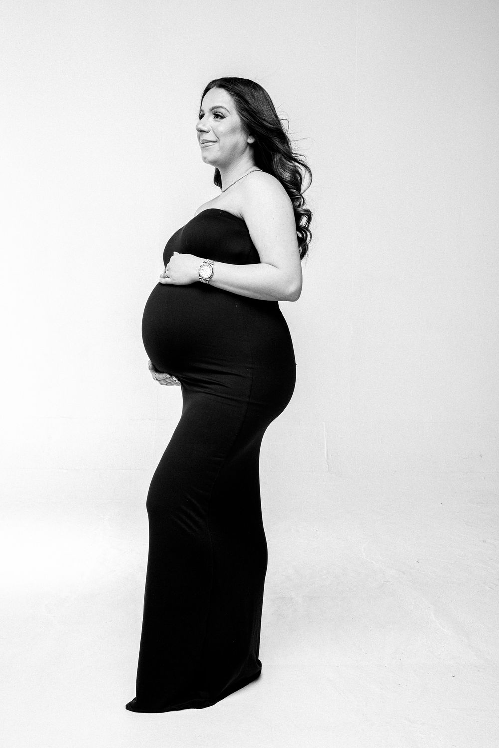 Elegant maternity portrait of a pregnant figure in a black strapless gown against a white backdrop.