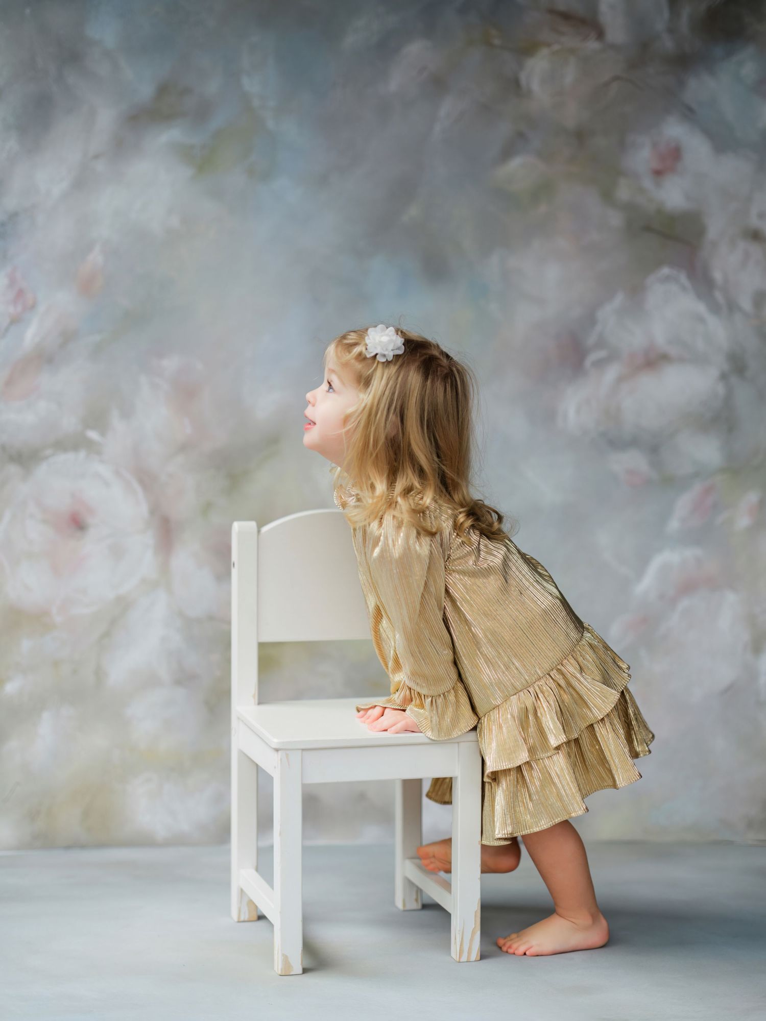 A small child in a ruffled beige dress leans playfully on a white chair against a dreamy painted backdrop.