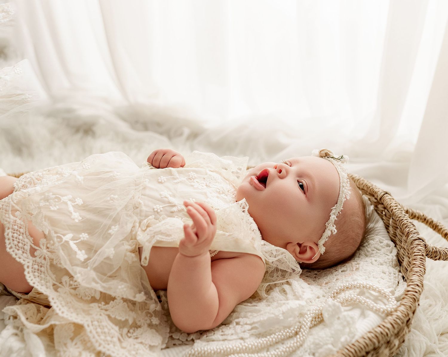 A baby wearing a lacy white christening gown and headband lies peacefully on soft white fabric.