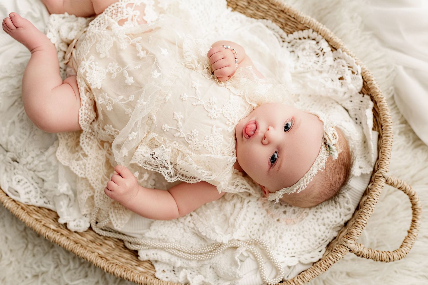 Small infant wearing delicate white lace dress and headband lying in woven basket on soft fabric.
