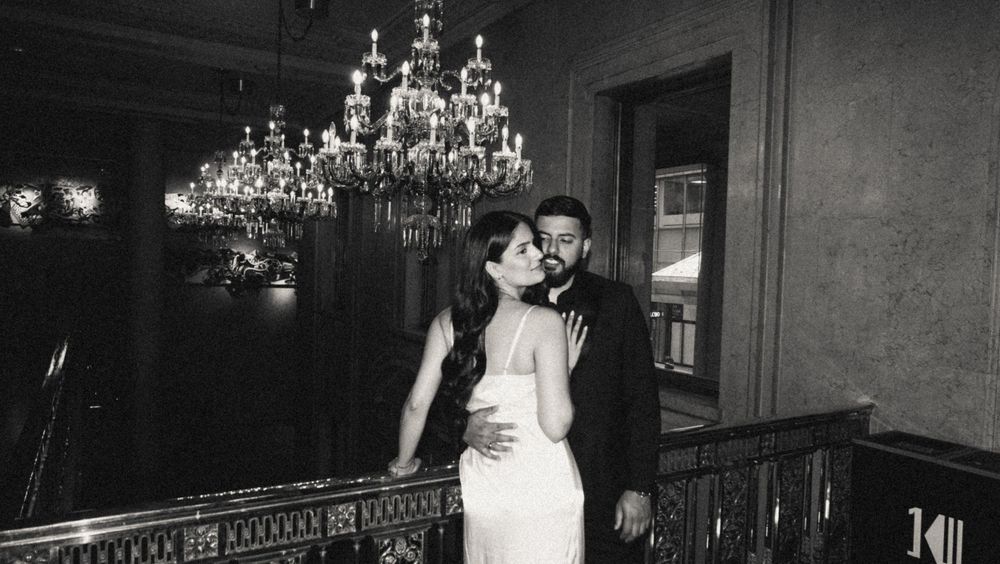 A romantic vintage black and white photo of a couple embracing under elegant crystal chandeliers in a formal ballroom setting.