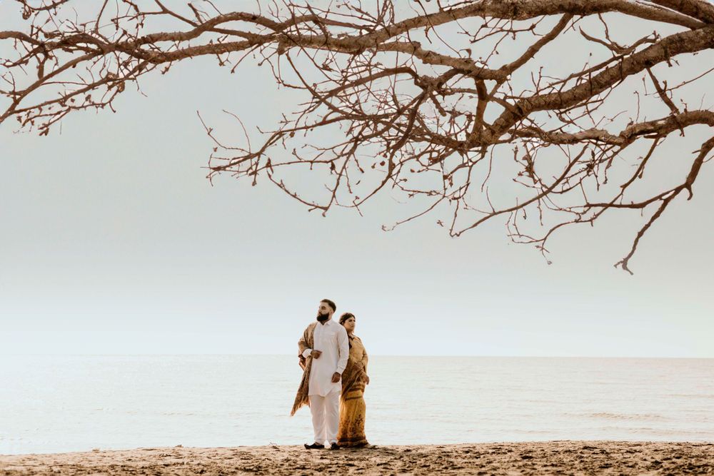 Two figures share a tender moment on a beach at sunset under bare tree branches stretching overhead.