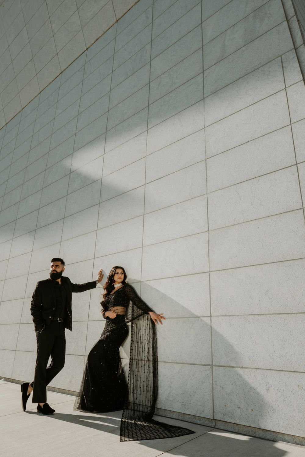 Two figures in dark formal attire pose dramatically against a modern white geometric wall with angular shadows.