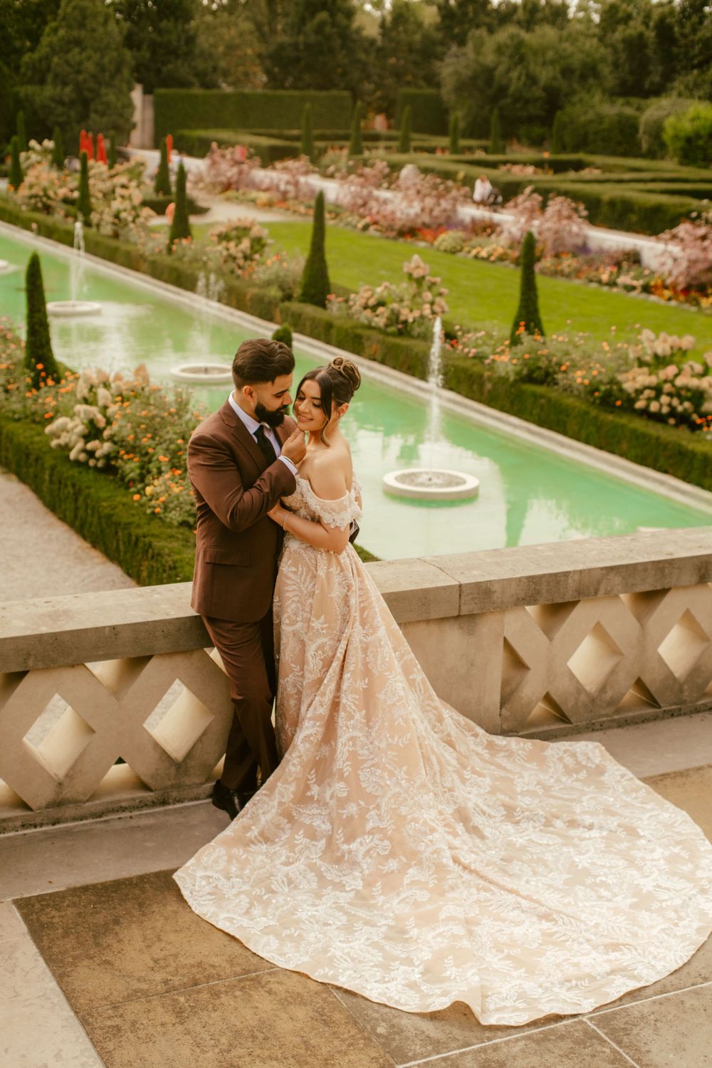 A romantic wedding couple embraces by an elegant garden fountain surrounded by manicured hedges and blooming flowers.