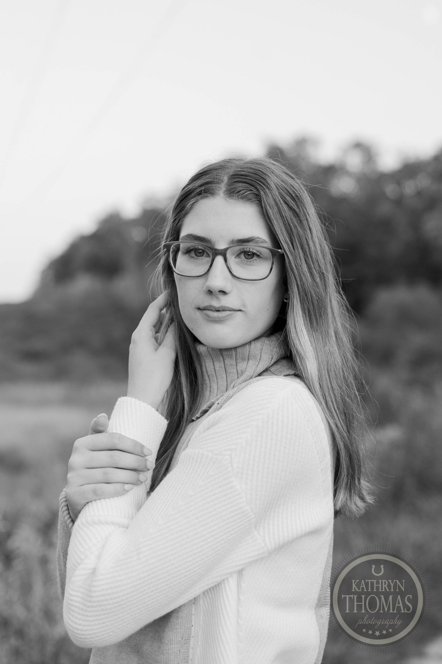Black and white portrait of someone in glasses and a white sweater against a blurred natural background.