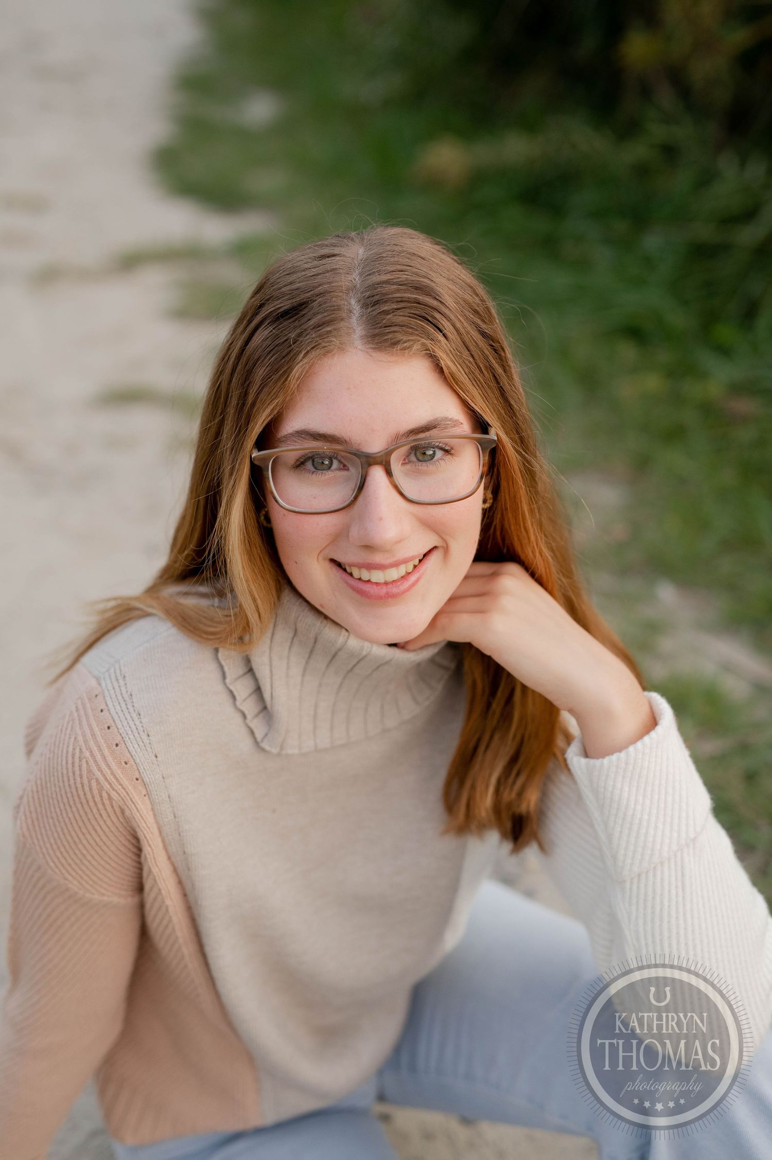 Natural light portrait wearing glasses and beige turtleneck against blurred outdoor backdrop.