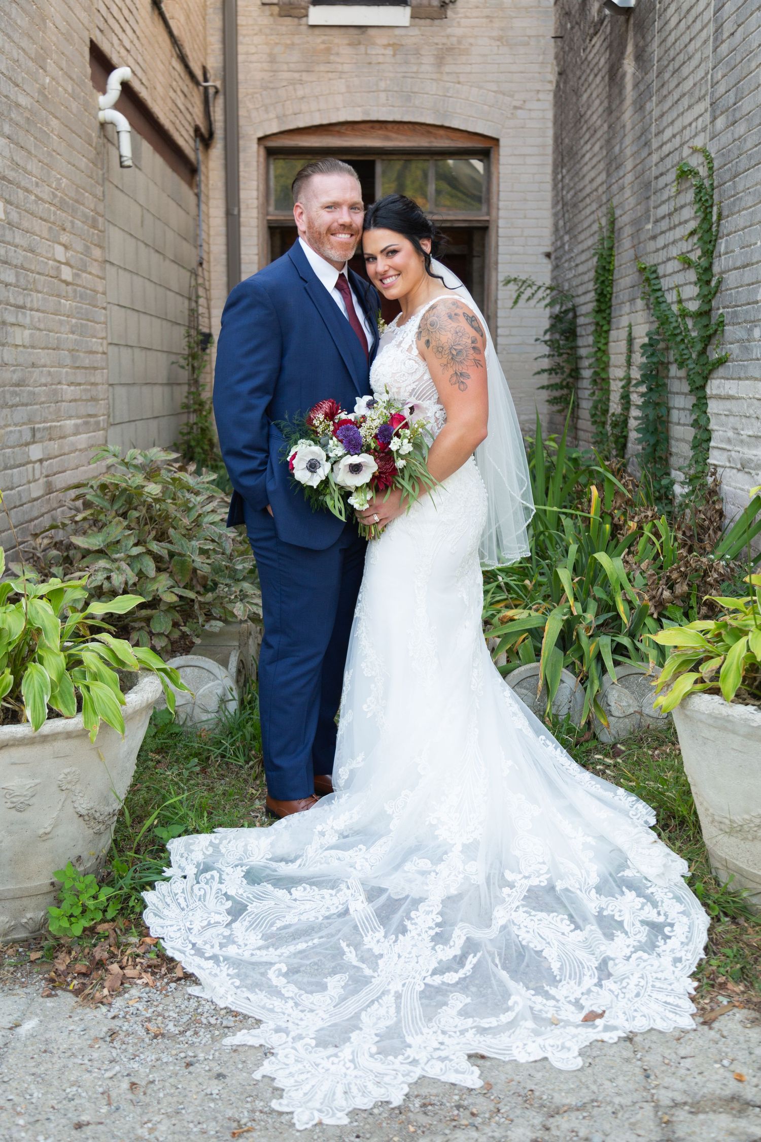 Wedding couple poses outdoors with lush greenery and brick backdrop, bride wearing lace train gown and holding bouquet.