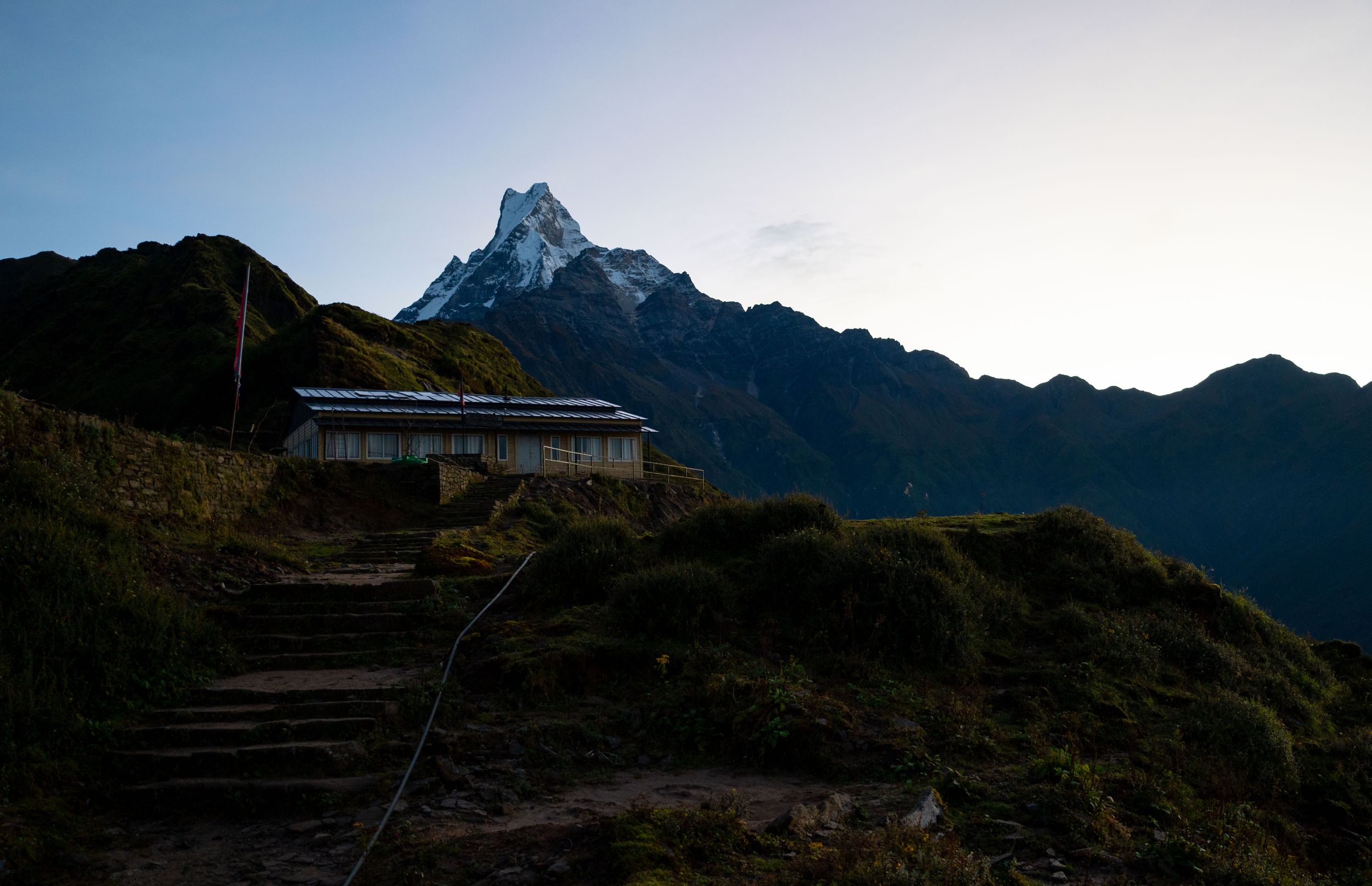 Mountain lodge nestled on hillside with dramatic peaks rising in background under blue sky.