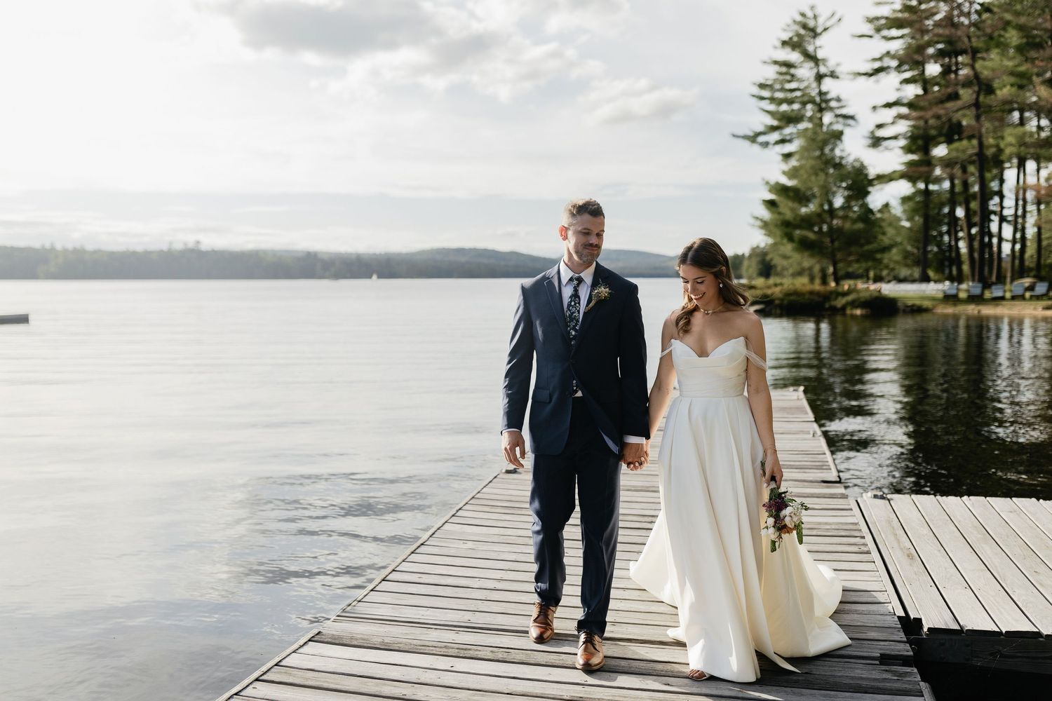 Two newlyweds walk hand in hand on a wooden dock overlooking a serene lake surrounded by pine trees.