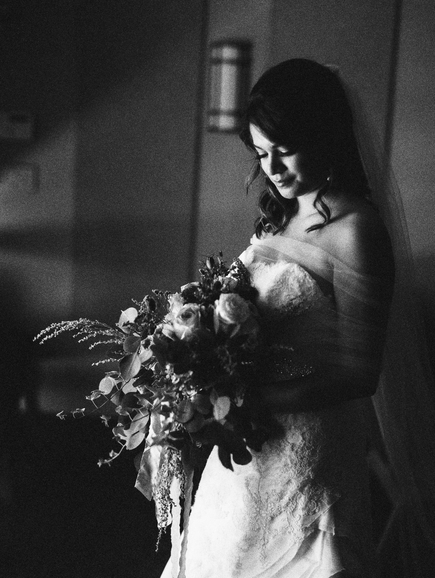 A bride in a white lace wedding dress gazes down at her elegant cascading bouquet in this black and white photograph.