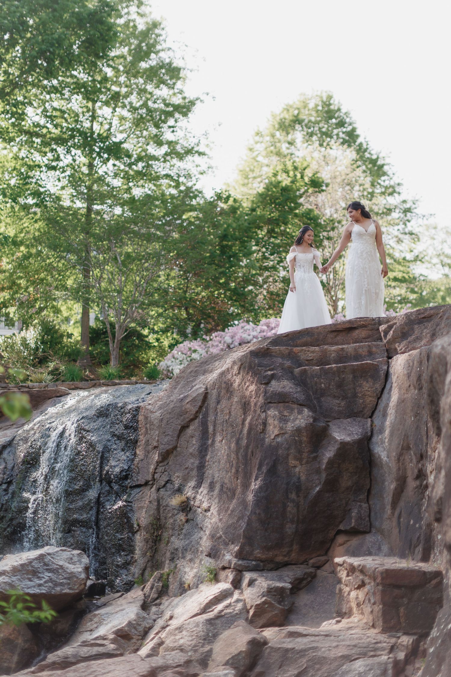 Two people in white dresses stand atop a rocky waterfall formation surrounded by lush green trees on a sunny day.