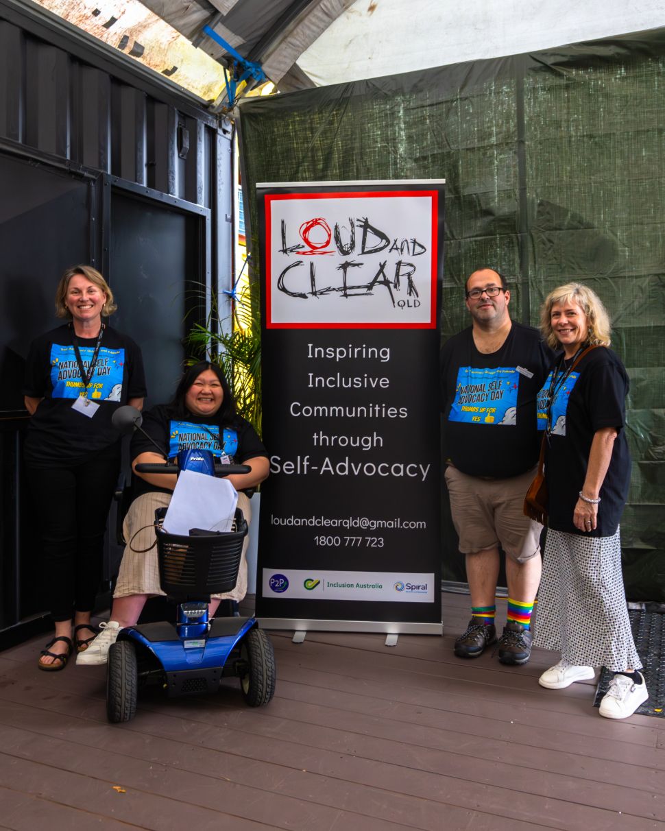 four people, three standing and one in a electric scooter chair with a loud and clear banner between them.