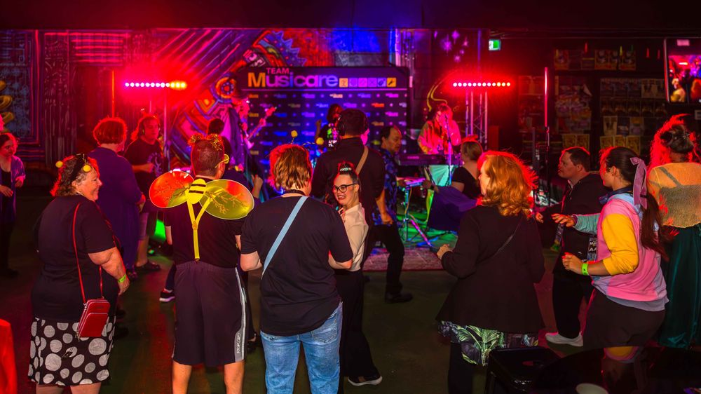 A low light, night seen with several people around a stage listening to music. Red and blue coloured lights in the background
