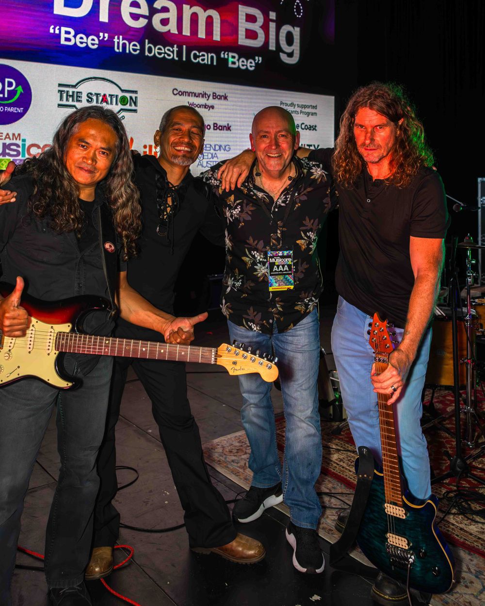 A four man musical band huddled together with there instruments stage side. Large digital display in the background