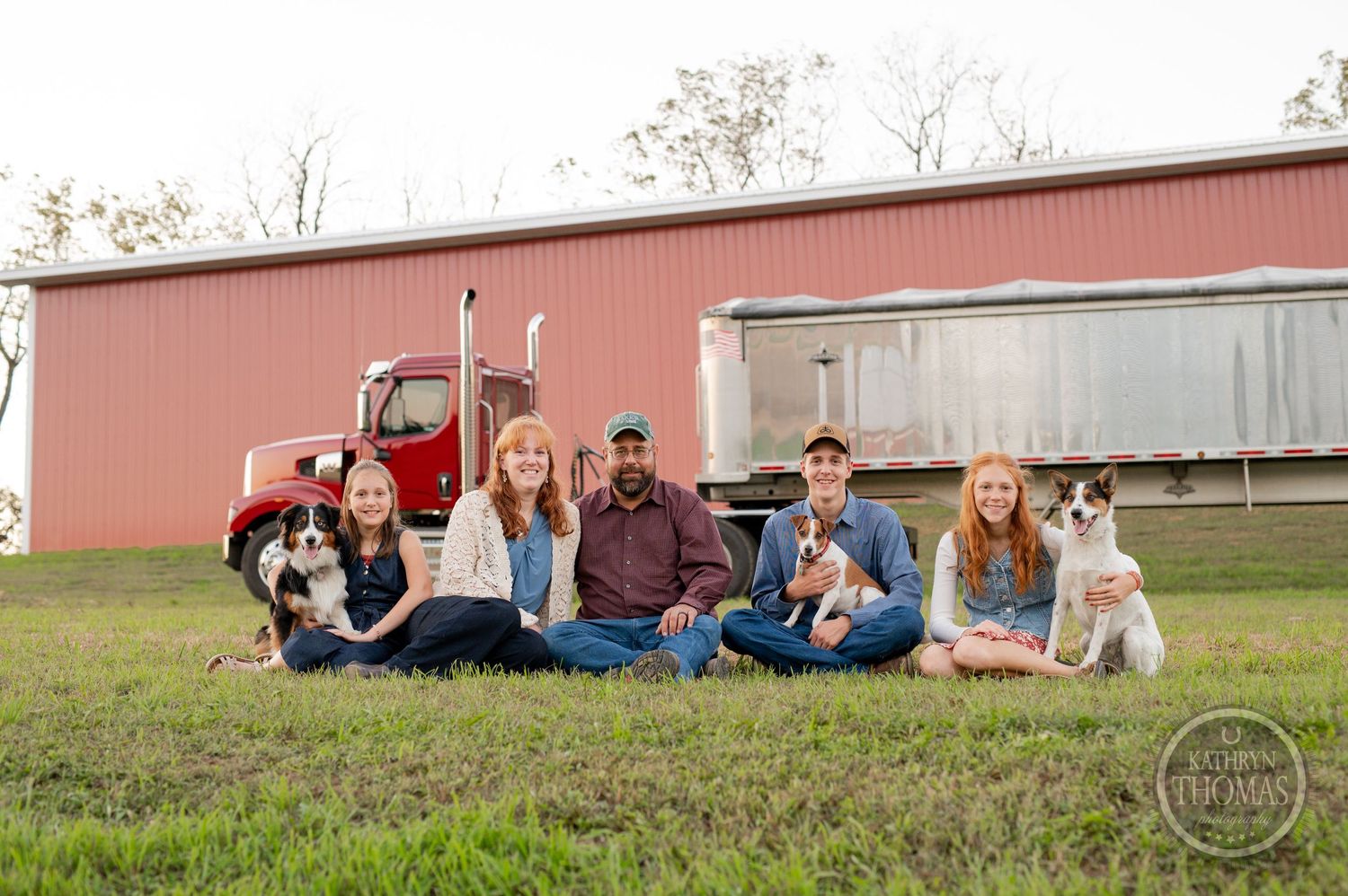 Family and dogs sitting together on grass with red barn and semi truck in background.