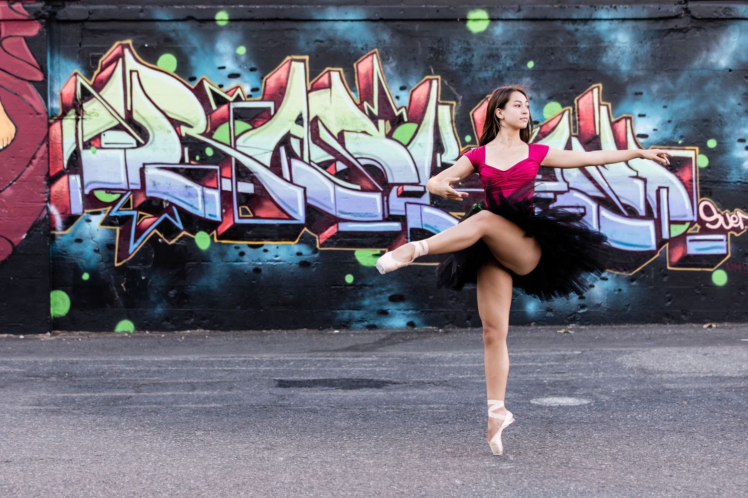 Ballet dancer in red dress strikes graceful pose en pointe against colorful graffiti wall backdrop in urban setting.