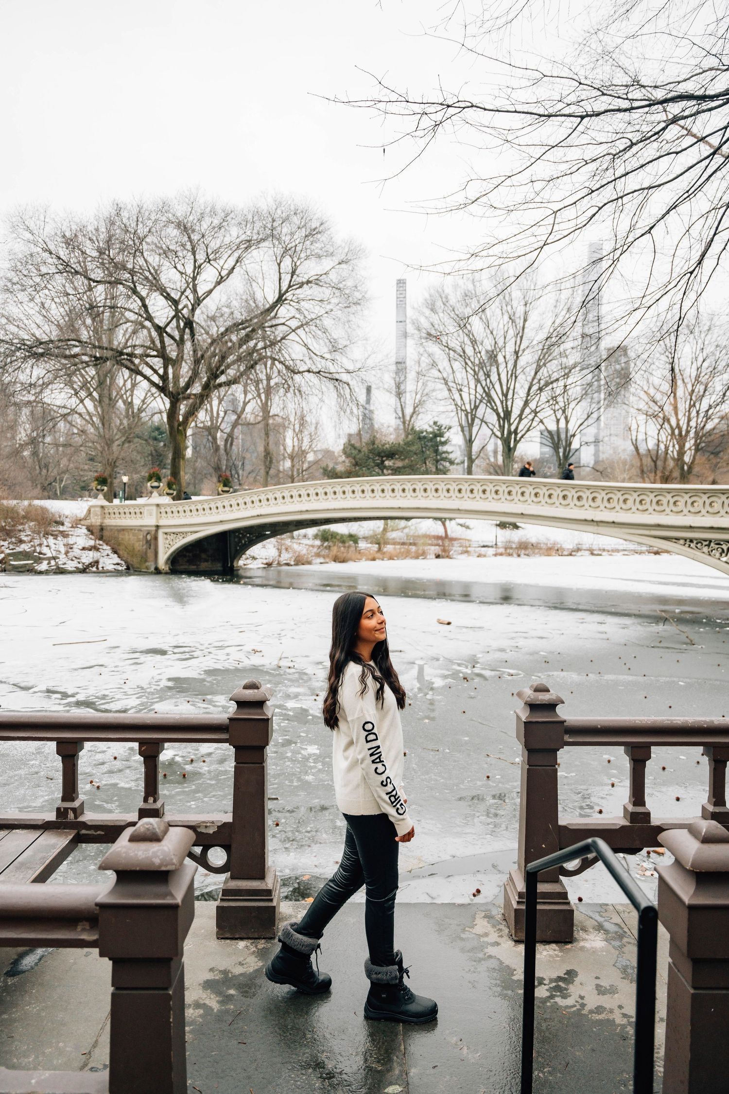 Winter scene at Bow Bridge in Central Park with person posing on snowy path.