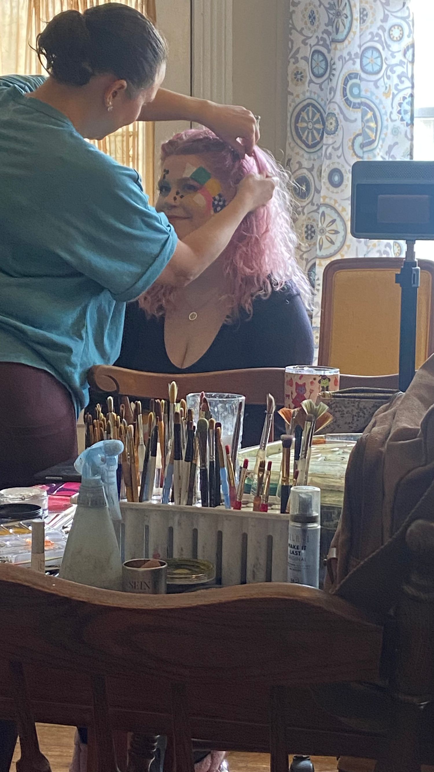 Professional hair stylist works on a client with bright pink hair while makeup supplies and brushes sit on wooden table.