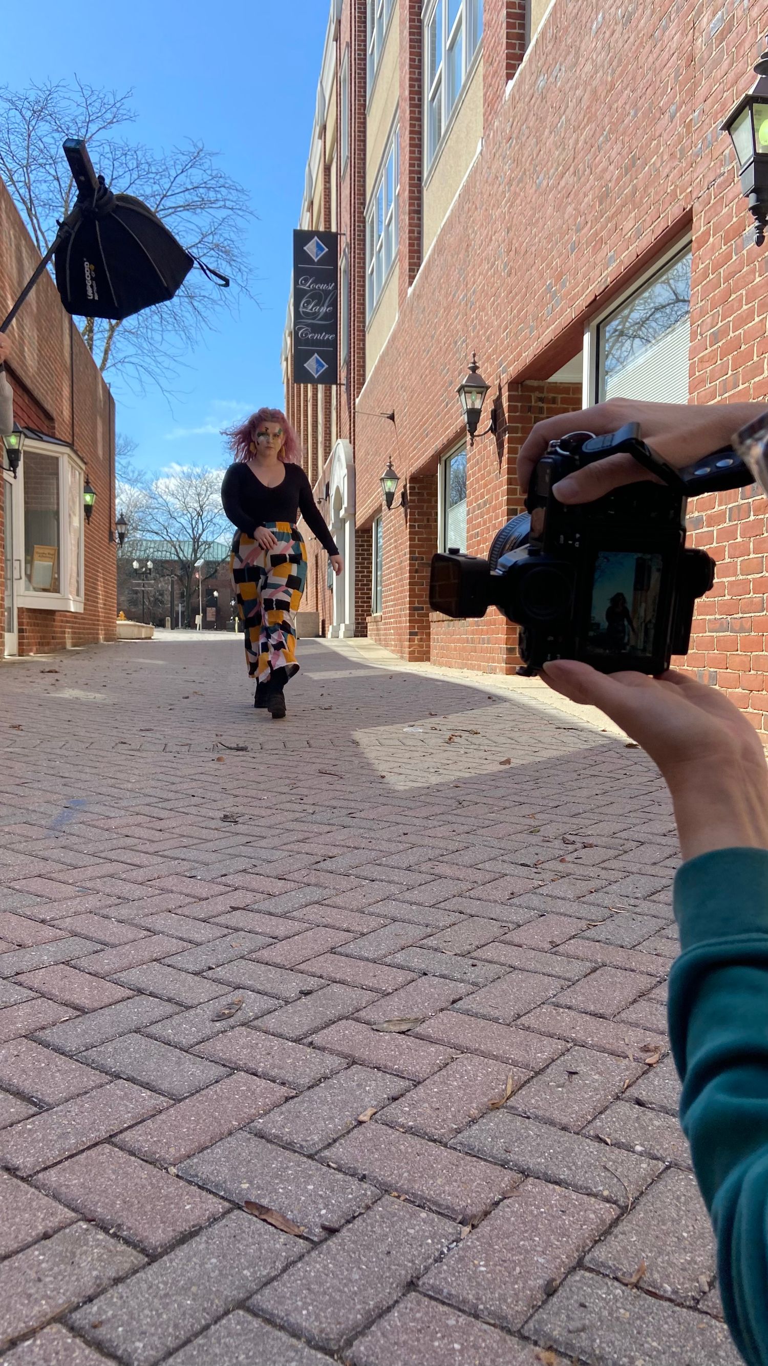 Someone taking a photo of a person in colorful patterned pants walking on a brick sidewalk between brick buildings.