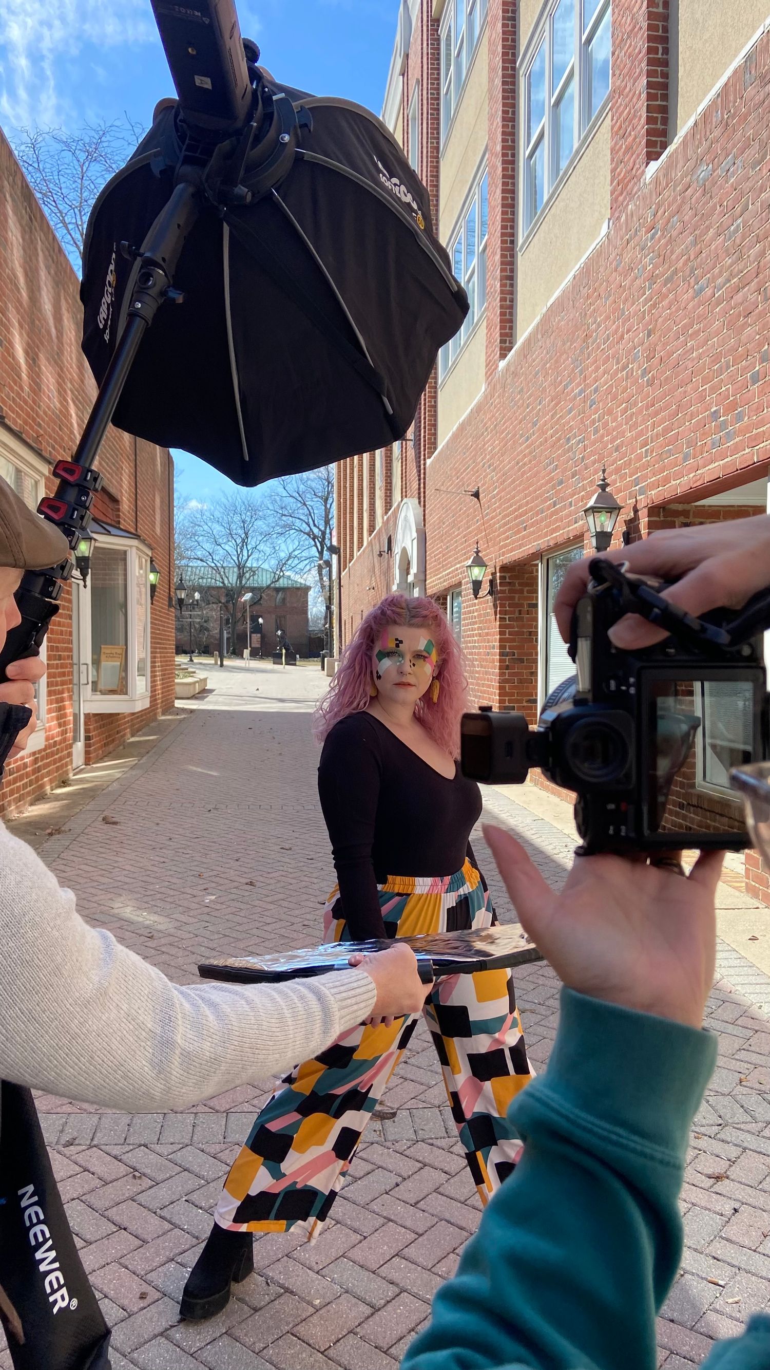 Behind-the-scenes video shoot on brick sidewalk with camera operator filming person in colorful patterned pants.