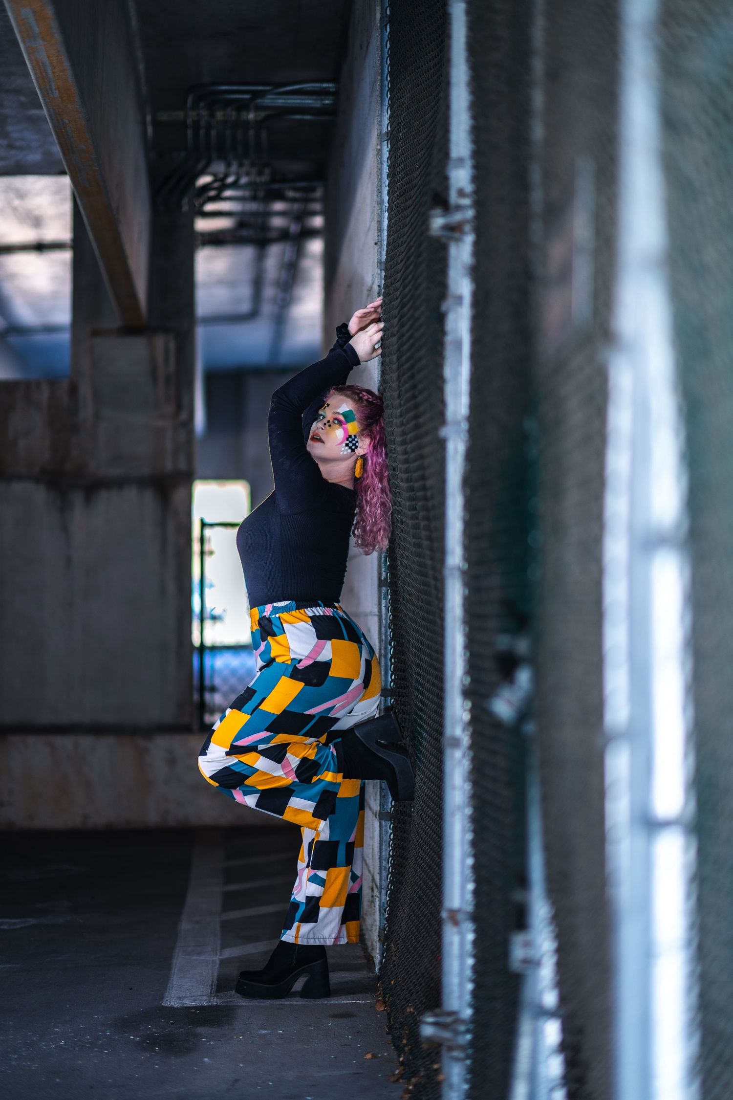 Street art performer wearing colorful geometric patterned pants and black top poses in urban underpass setting.