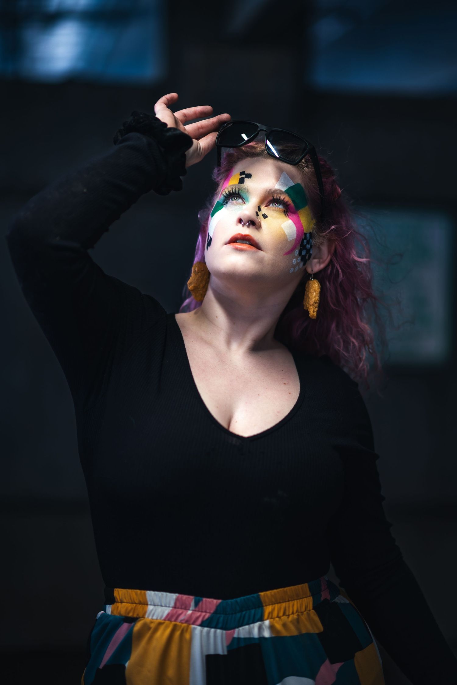 Creative portrait with vibrant rainbow eye makeup and colorful hair wearing black top and patterned skirt in moody lighting.