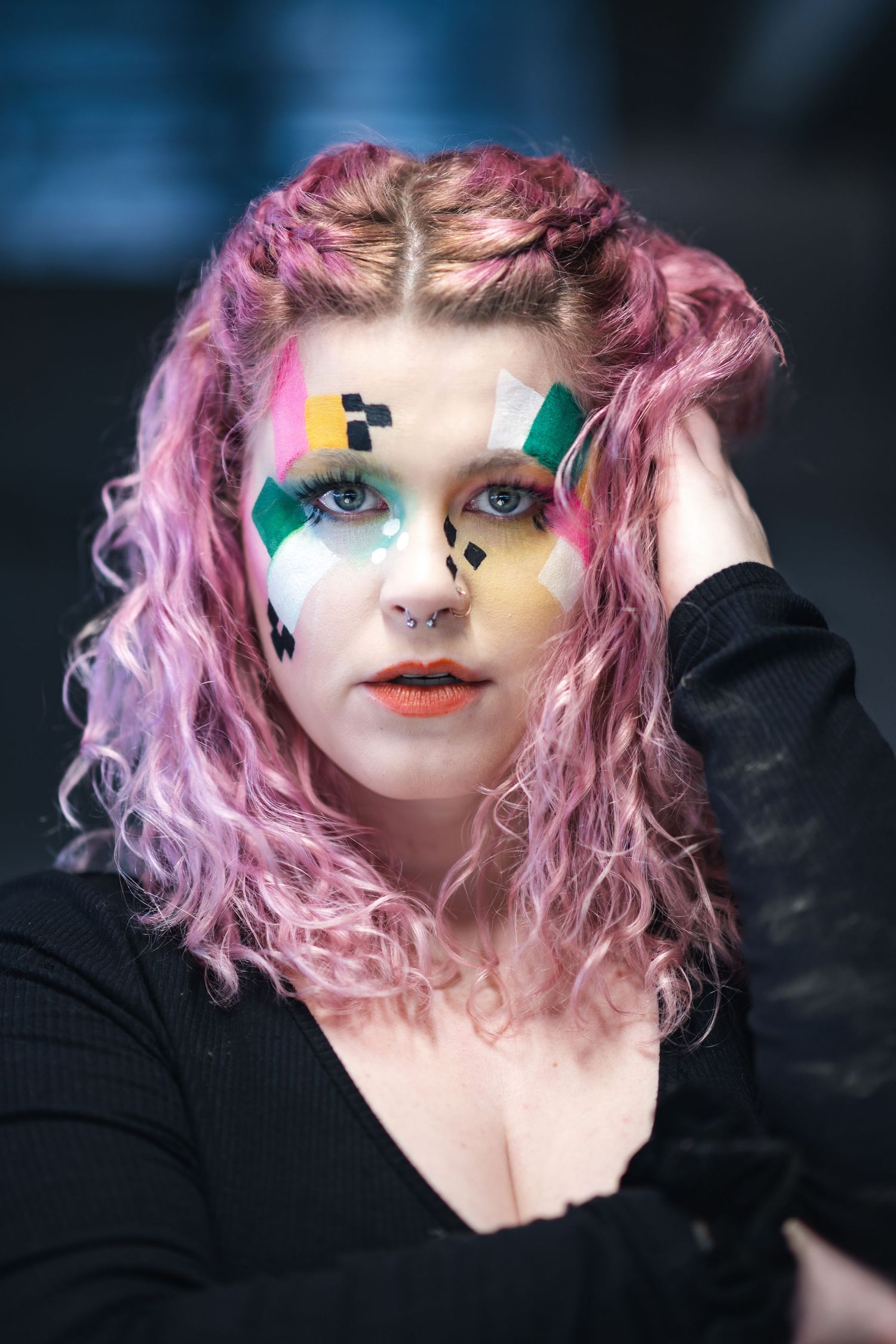 Creative makeup art featuring pink curly hair, geometric face paint designs in green and yellow, and coral lipstick.