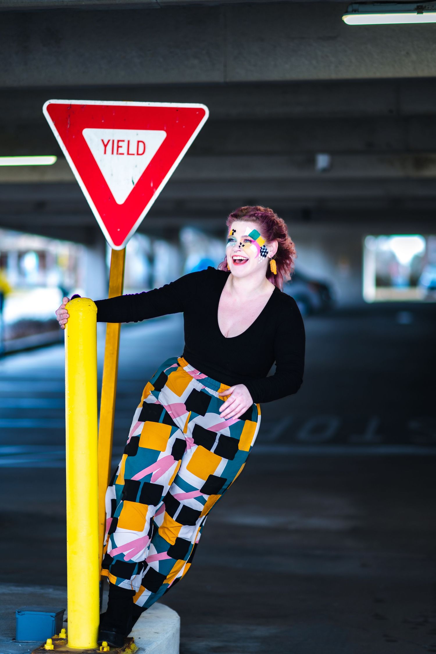 A figure in dramatic makeup wearing a black top and colorful geometric patterned pants poses by a yield sign in a parking garage.