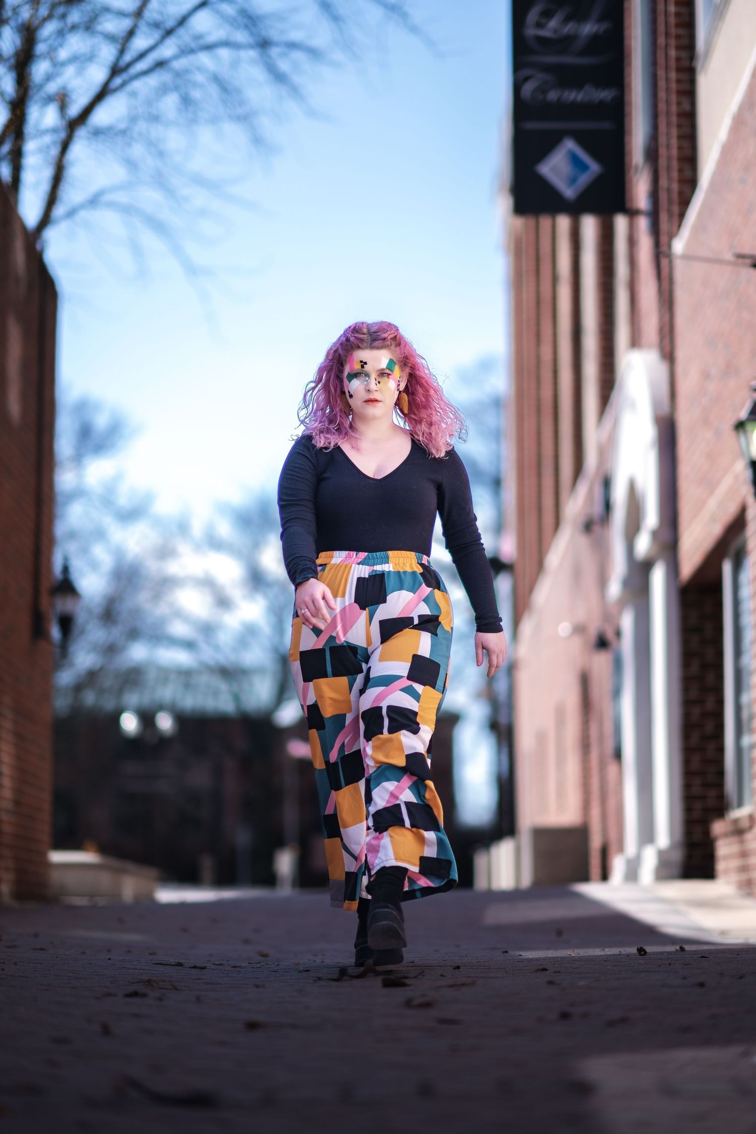 Someone in a black top and colorful patterned skirt walks down an alley with pink hair and dramatic makeup.