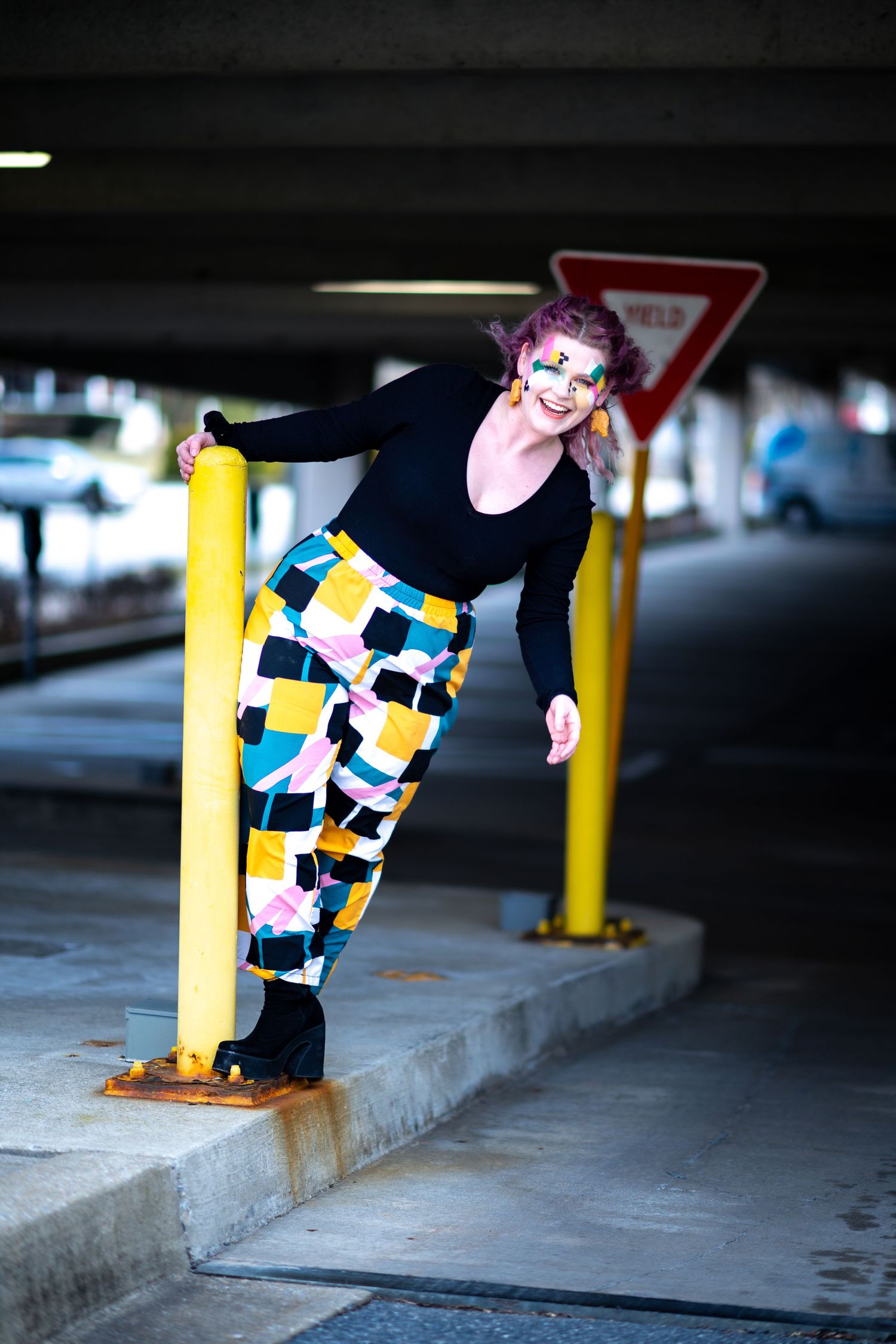 A person in colorful geometric patterned pants and black top poses playfully near yellow posts in a parking garage.