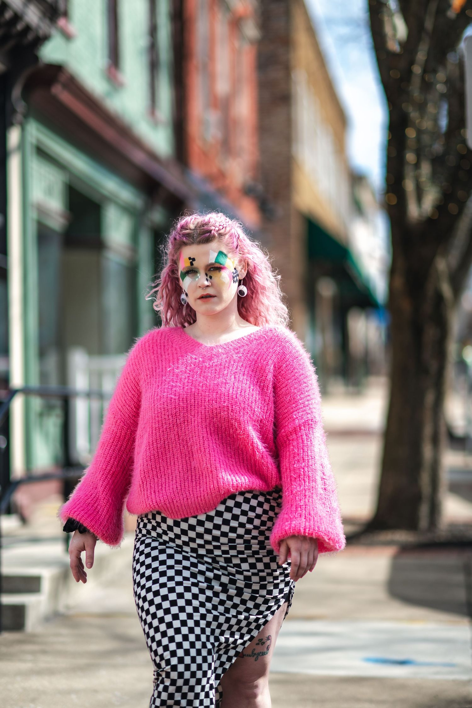 Person in bright pink sweater and checkered skirt poses on city sidewalk with storefronts in background.