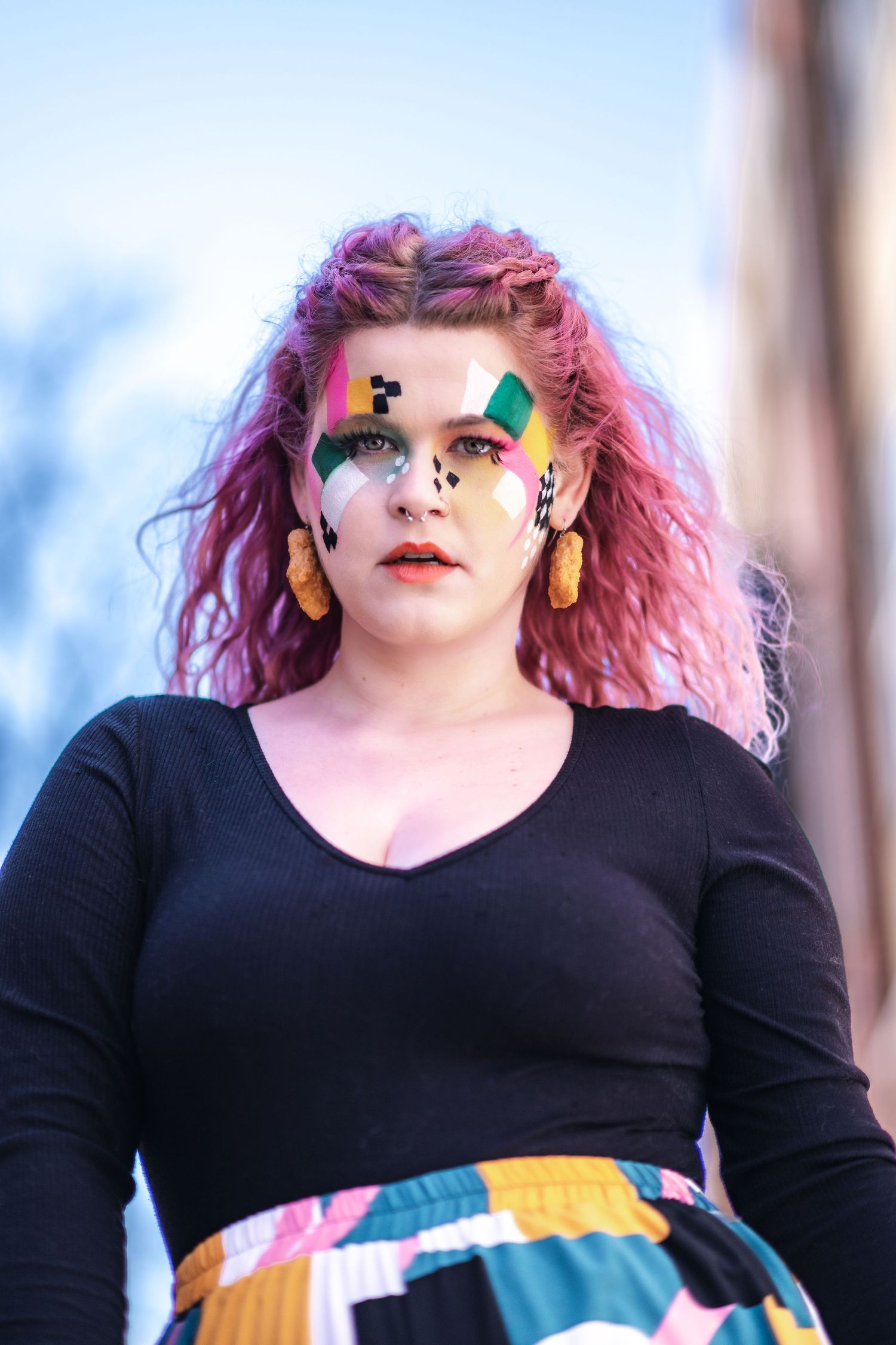 Person with bright pink hair wearing face gems, yellow earrings and colorful makeup against a blurred outdoor background.