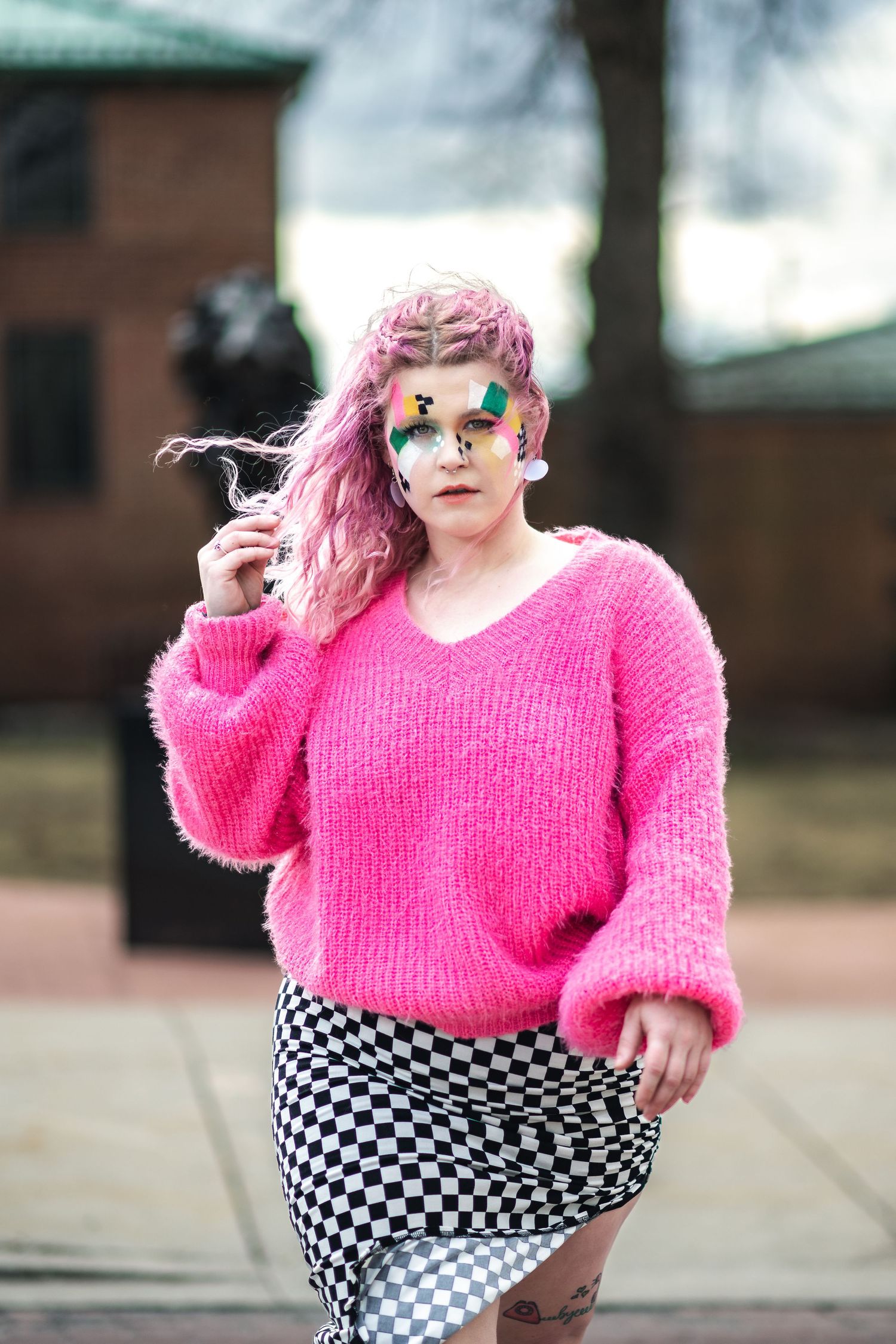 A person in a bright pink sweater and checkered skirt strikes a pose outdoors with dyed pink hair and artistic makeup.