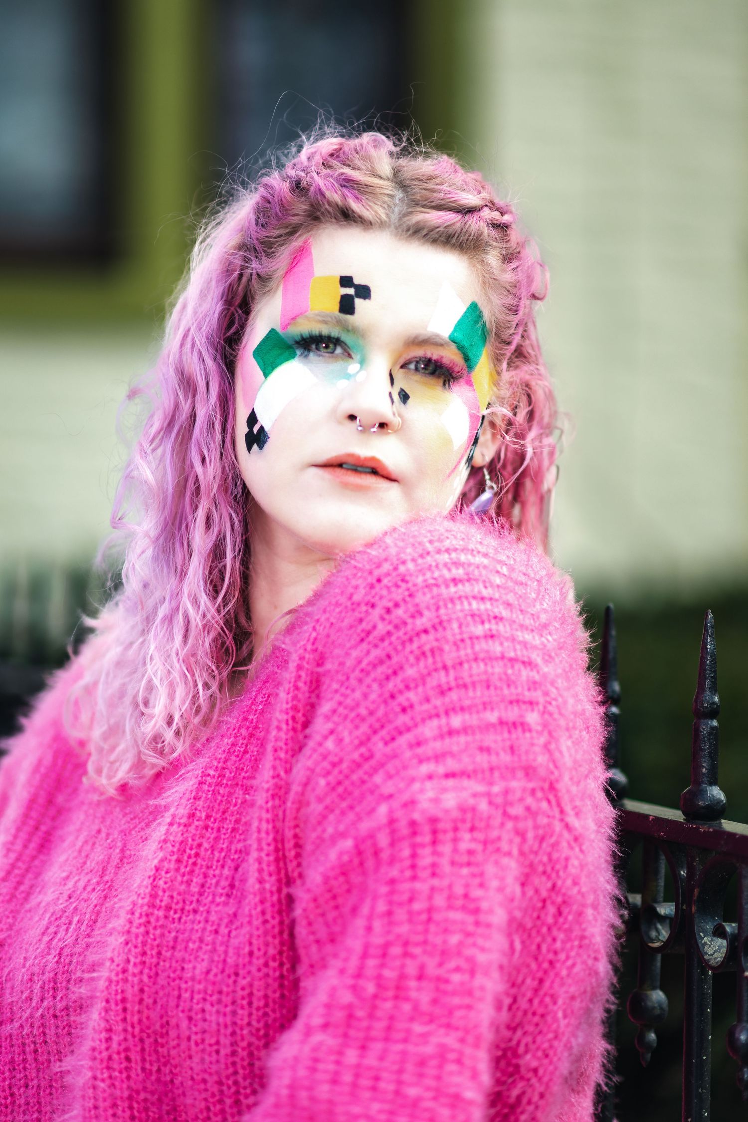 Creative portrait featuring bright pink hair and colorful face paint designs, wearing a vibrant pink knit sweater.