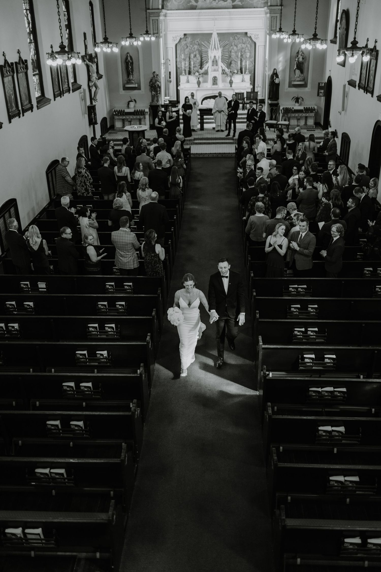 A black and white photo showing a couple walking down the church aisle after their wedding ceremony with guests seated in pews.
