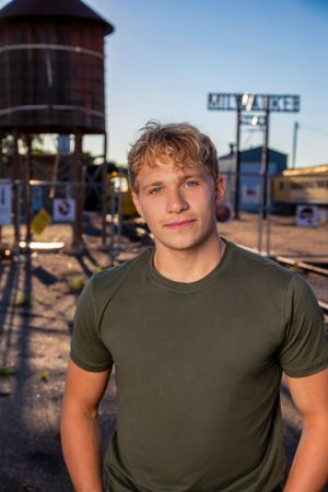 A person in an olive green t-shirt stands in front of a rustic water tower and railroad crossing sign.