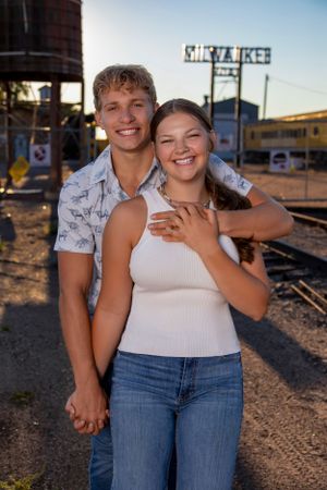 A happy couple poses together outdoors at sunset near train tracks and old industrial buildings.