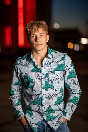 Portrait of someone wearing a stylish white and green patterned button-up shirt at night with city lights in background.