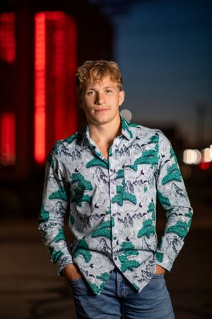 A person poses outdoors at night wearing a trendy green and white patterned button-up shirt with jeans.