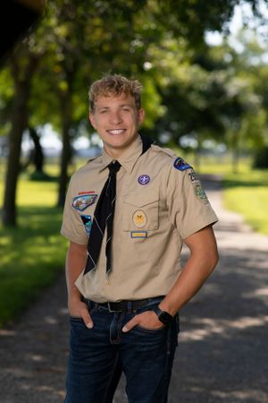 A Scout in tan uniform with patches and black tie standing on a park path with trees in the background.