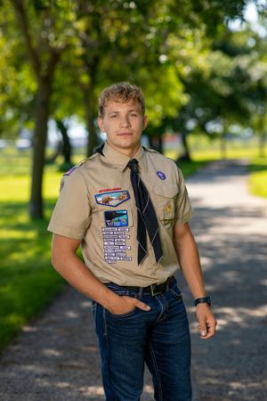 Scout in tan uniform with badges and patches stands on tree-lined path in a park on sunny day.